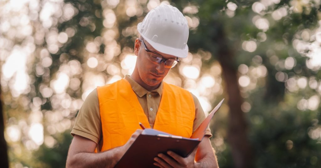 An arborist in a high-vis orange vest and white hard hat writes information on a clipboard as they stand near trees.