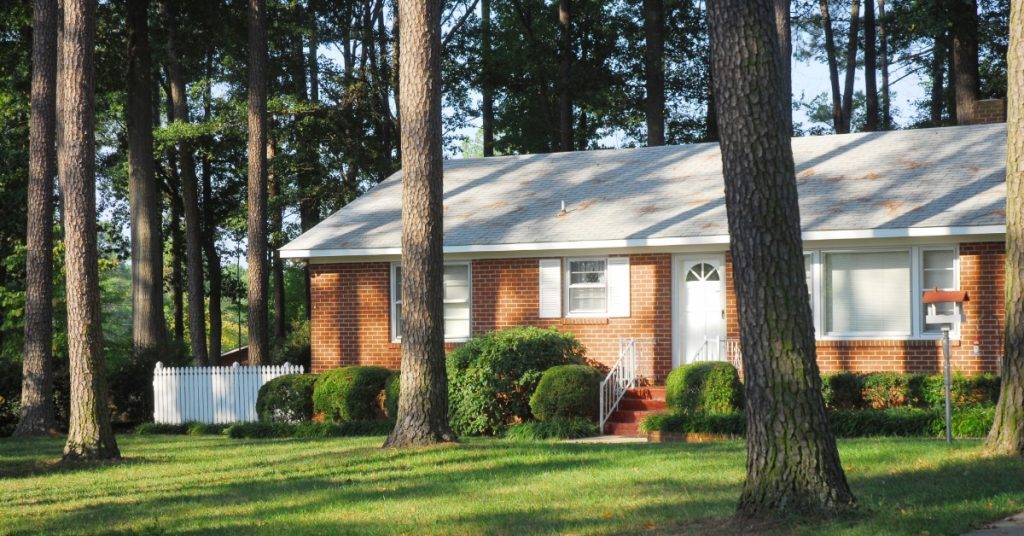 A brick home sits facing the sunset. The front yard is freshly groomed with mature trees scattered around.