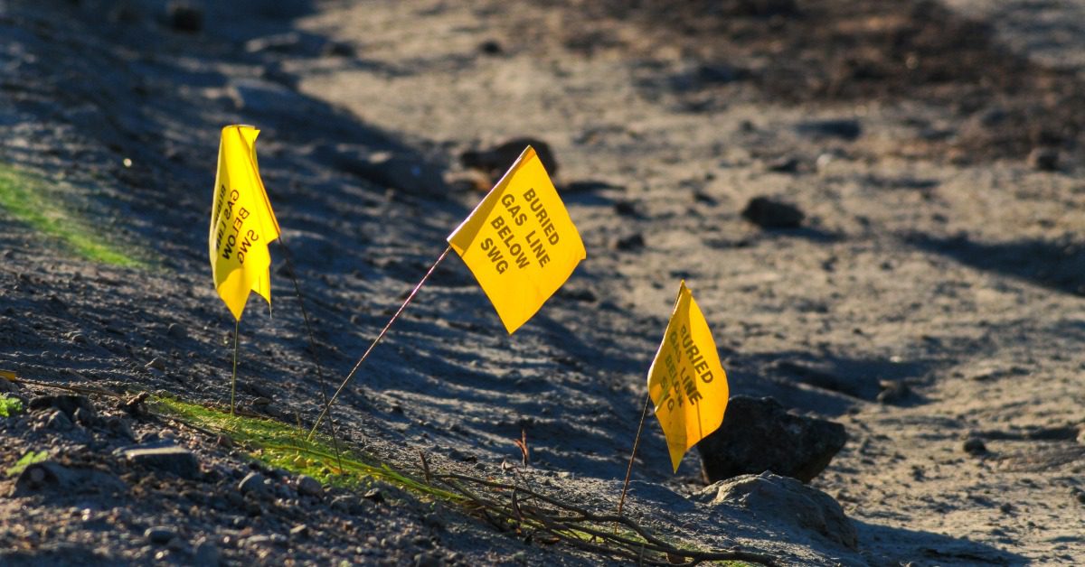 Three yellow locate flags in the ground on a dirt path signaling gas, oil, or petroleum underground.