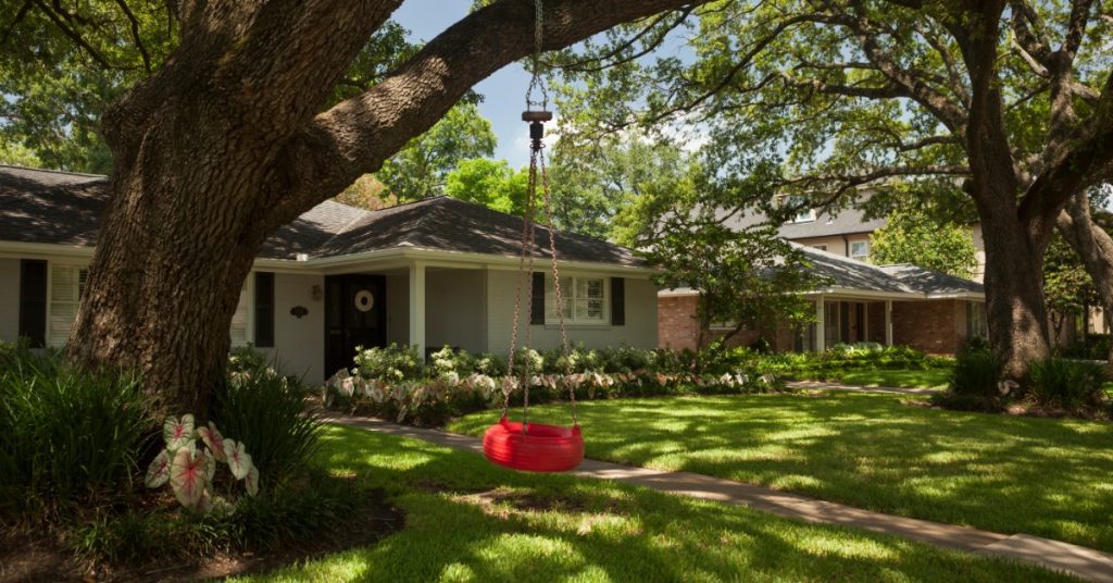 A bright red tire swing hangs from a large tree branch. The tree is old and sits close to the residential property.